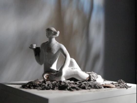 A white porcelain figurine sits on a pile of ash.